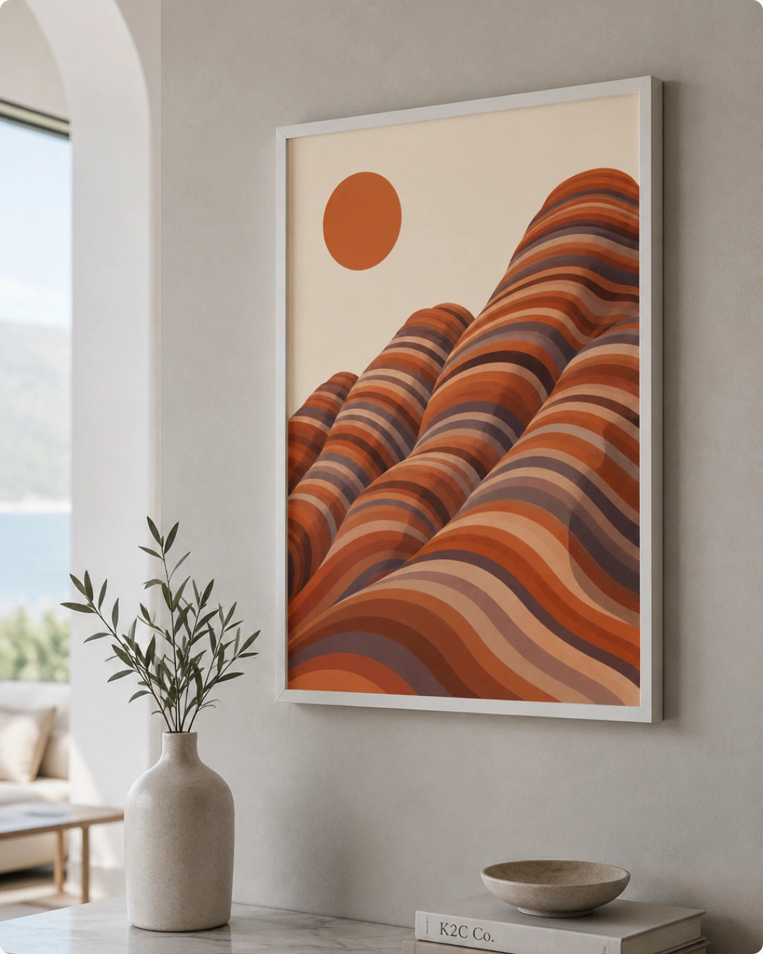 Framed abstract landscape wall art hanging on a light gray wall in a styled interior. The artwork shows layered wavy hills in terracotta, rust, orange, and brown tones with a muted orange sun on a warm cream background.