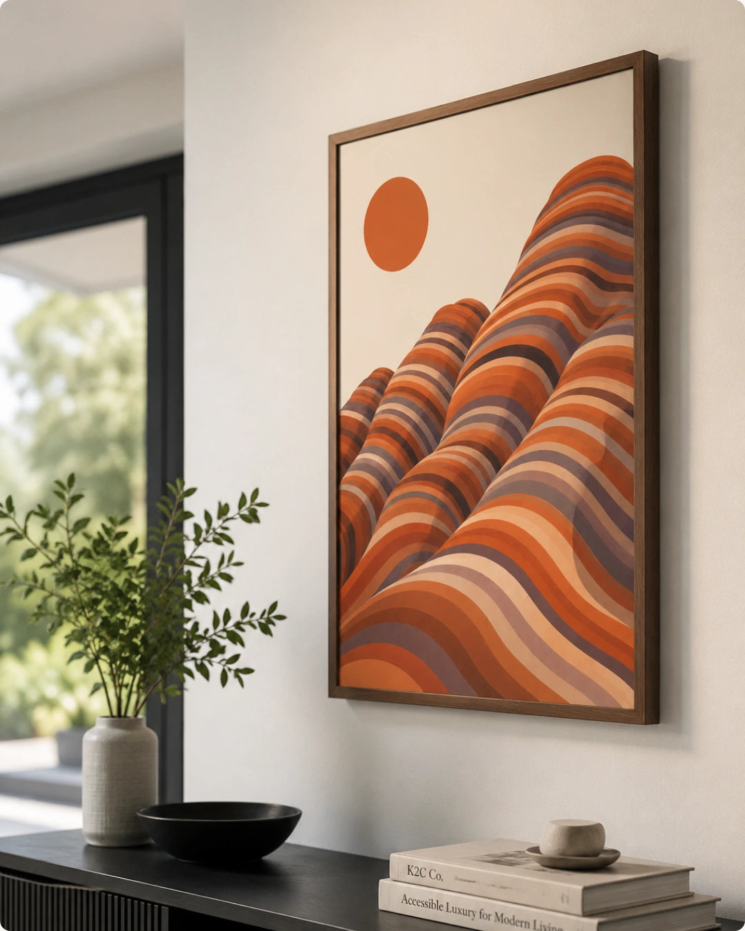 A framed abstract landscape art print featuring wavy orange, terracotta, and brown hills under a solid orange sun on a cream background, styled on a white wall above a console table with books and a plant.