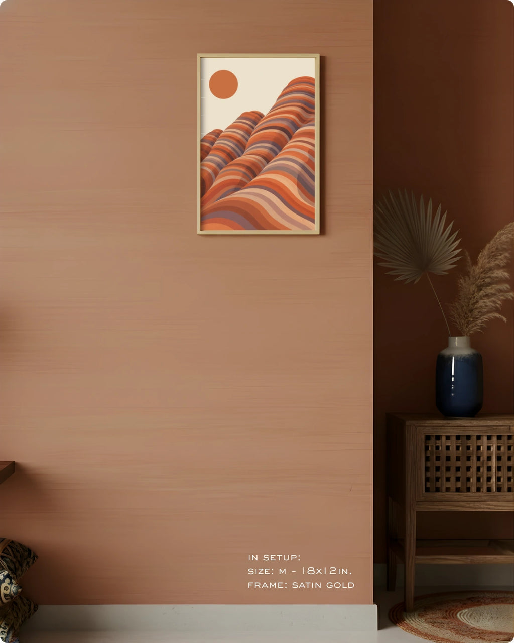 A framed wall art print hangs on a warm terracotta wall in a styled interior. The artwork shows layered wavy hills in orange, rust, beige, and muted blue tones with a small orange sun.