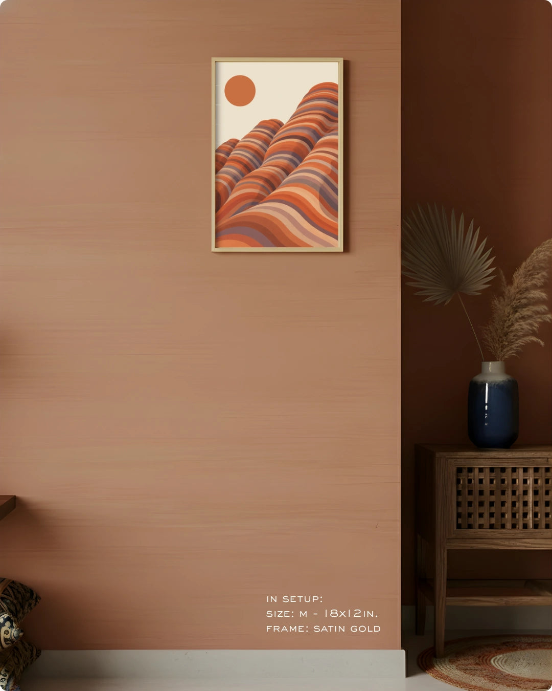 A framed wall art print hangs on a warm terracotta wall in a styled interior. The artwork shows layered wavy hills in orange, rust, beige, and muted blue tones with a small orange sun.