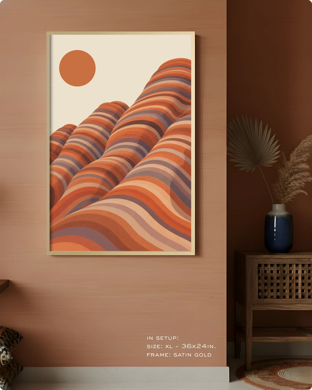 A framed wall art print featuring abstract wavy desert-like hills in orange, terracotta, and beige tones, hung on a warm brown wall in a styled interior with a side table and vase.