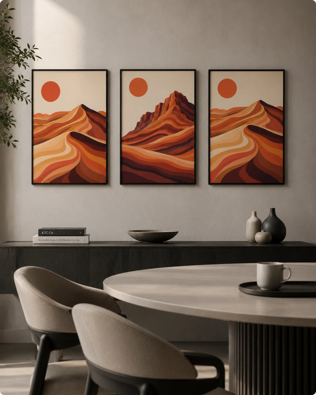 A three-panel framed wall art set hangs above a dark console table in a modern dining room. Each panel shows an abstract desert landscape with layered orange and brown hills under a muted orange sun on a cream background.