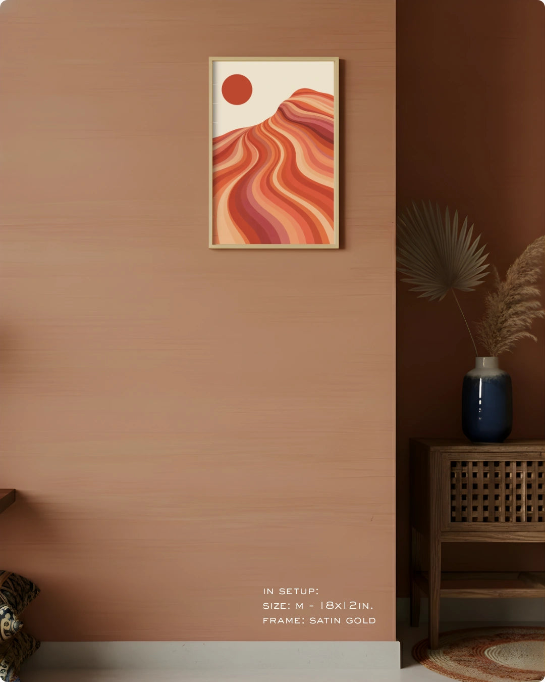 A framed abstract wall art print hangs on a warm terracotta wall in a styled interior. The artwork features flowing layered hills in orange, red, and pink tones with a red sun on an off-white background, enclosed in a satin gold frame.