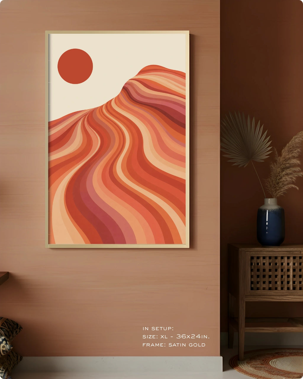 A framed abstract landscape art print featuring wavy orange, red, and peach hills under a solid orange sun on a cream background, displayed on a warm brown wall in a styled interior setting.