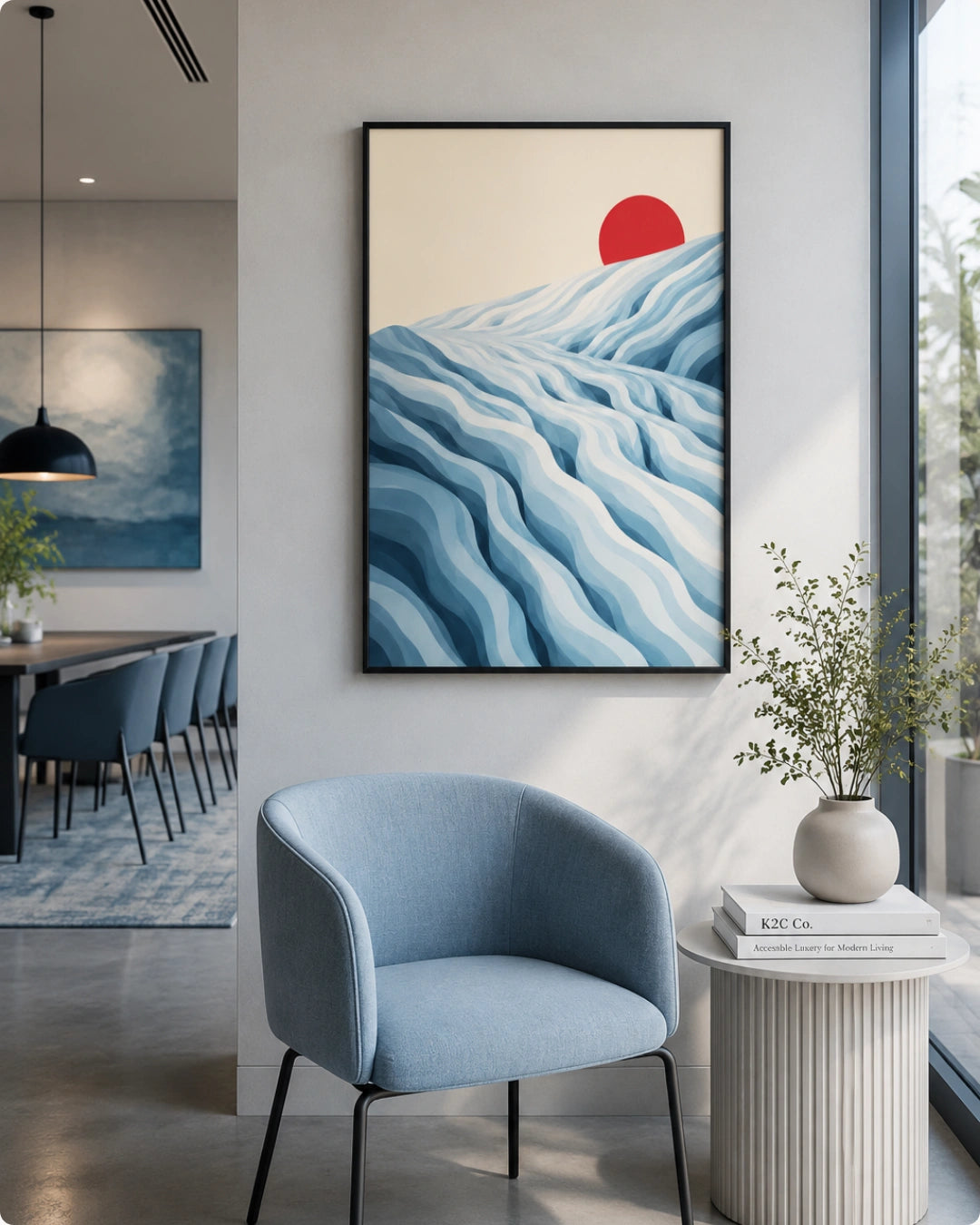 A framed abstract art print featuring blue wavy hills and a red sun on a cream background, displayed on a light gray wall in a modern living room setting with a blue armchair and a side table.