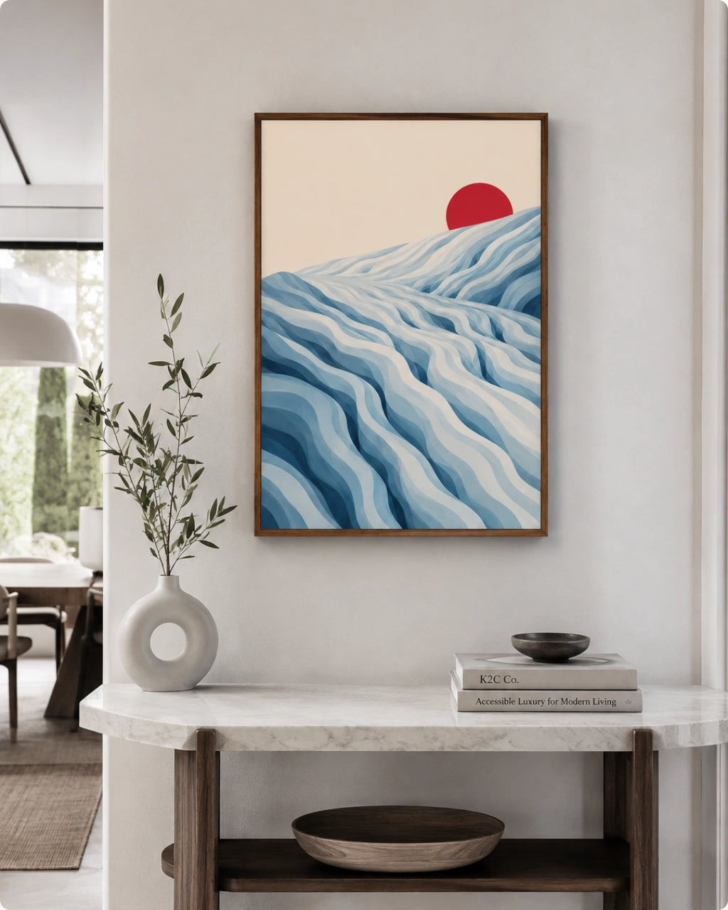A framed art print featuring a minimalist landscape with blue wavy hills and a red sun on a cream background, displayed on a white wall above a marble console table in a modern interior setting.