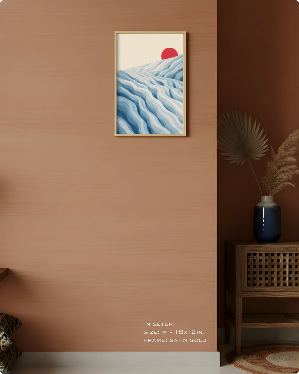A framed wall art print hangs on a warm terracotta wall in a styled interior. The artwork shows layered blue waves with a red sun on an off-white background, in a satin gold frame.