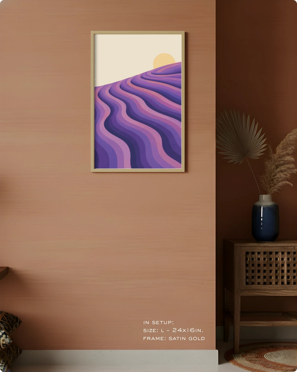 A framed art print featuring stylized purple and lavender wavy hills under a pale yellow sun, displayed on a light gray wall in a modern office setting with a desk, laptop, chair, and potted plant.