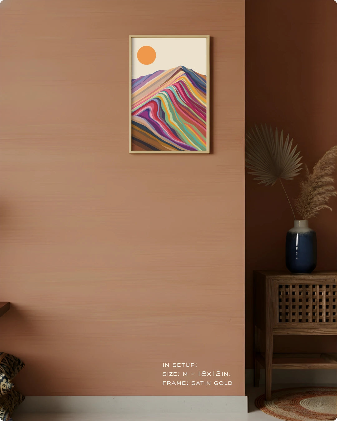 A framed abstract wall art print hangs on a warm terracotta wall in a styled interior. The artwork shows a multicolored mountain-like wave pattern with a small orange sun in the upper left.