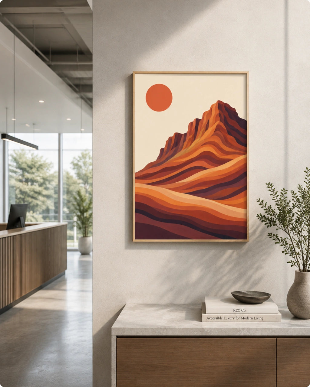 A framed art print featuring a stylized desert mountain landscape with a red sun, displayed on a light gray wall above a wooden desk in a modern, sunlit room.