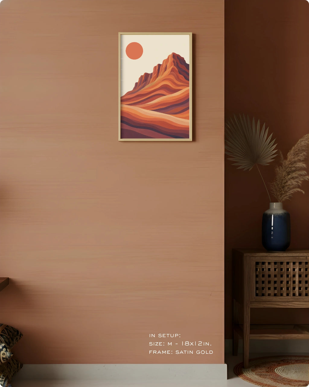 A framed art print featuring a stylized desert mountain landscape with a red sun, displayed on a light gray wall above a wooden desk in a modern, sunlit room.