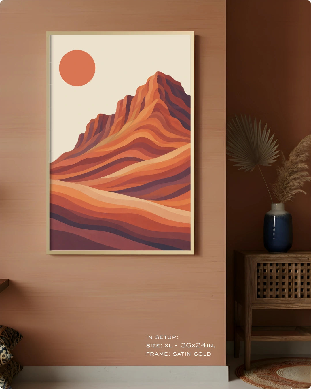 A framed desert landscape art print hangs on a warm brown wall in a styled interior. The print shows layered orange, red, and brown mountain or canyon shapes with a muted orange sun on a cream background.