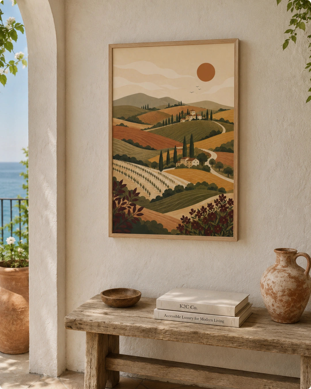A framed landscape art print featuring a Tuscany vineyard scene hangs on a textured cream wall above a rustic wooden bench with books and a terracotta jug. The print is shown in a styled home environment with a glimpse of the blue sea and sky on the left.