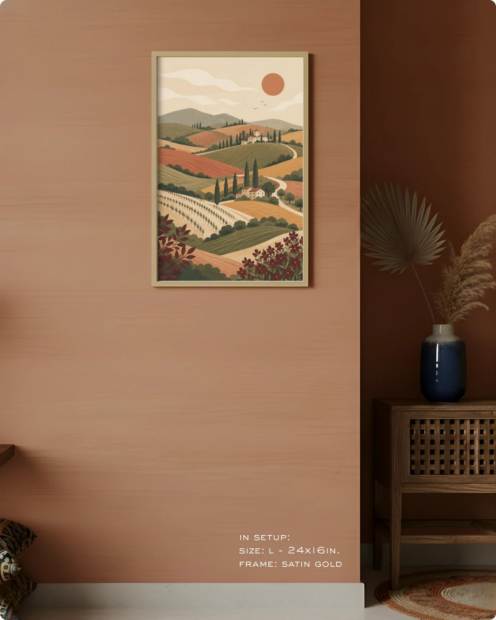 A framed landscape art print hangs on a warm terracotta-colored wall in a styled interior setting. The print depicts rolling hills, vineyards, cypress trees, and a sun in muted earthy tones.
