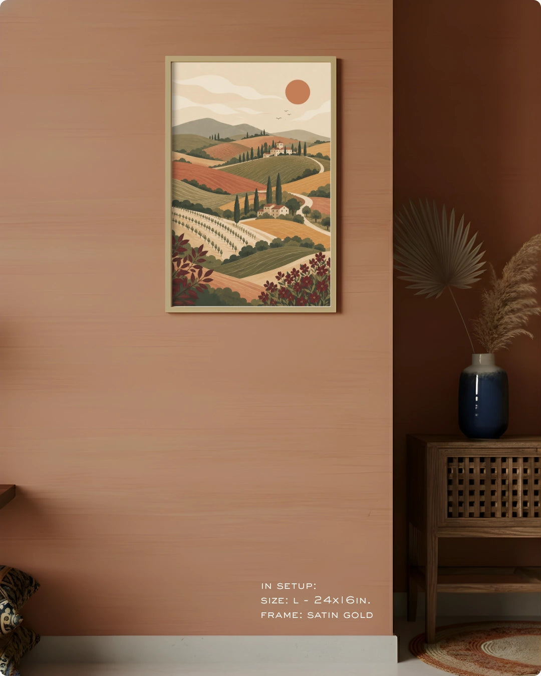A framed landscape art print hangs on a warm terracotta-colored wall in a styled interior setting. The print depicts rolling hills, vineyards, cypress trees, and a sun in muted earthy tones.