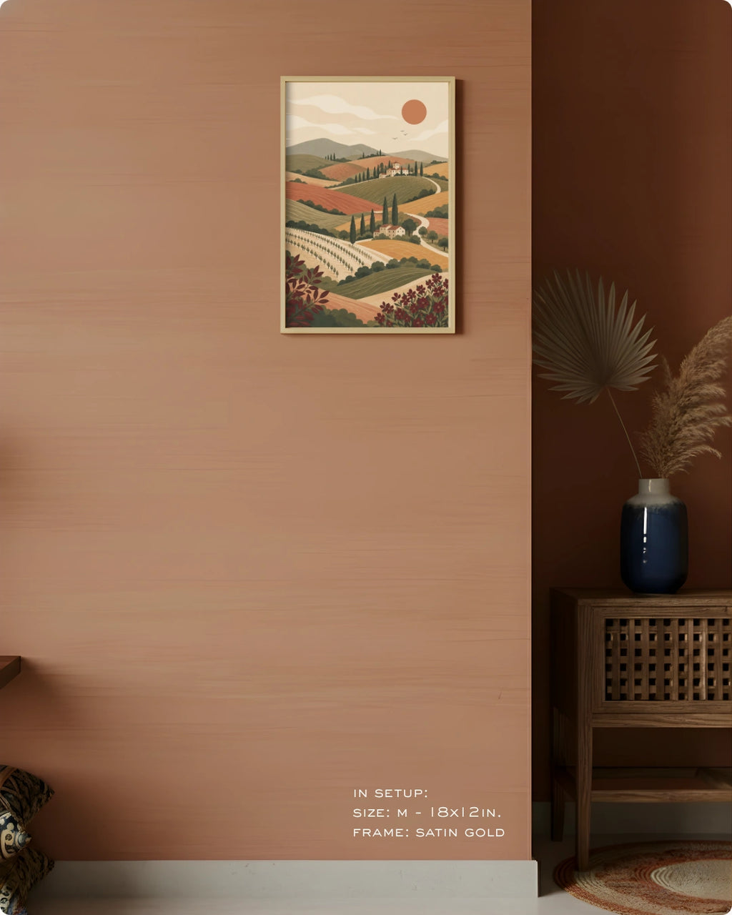 A framed landscape art print hangs on a terracotta-colored wall in a styled interior setting. The print depicts a stylized Tuscany vineyard with olive trees, rolling hills, and a sun.