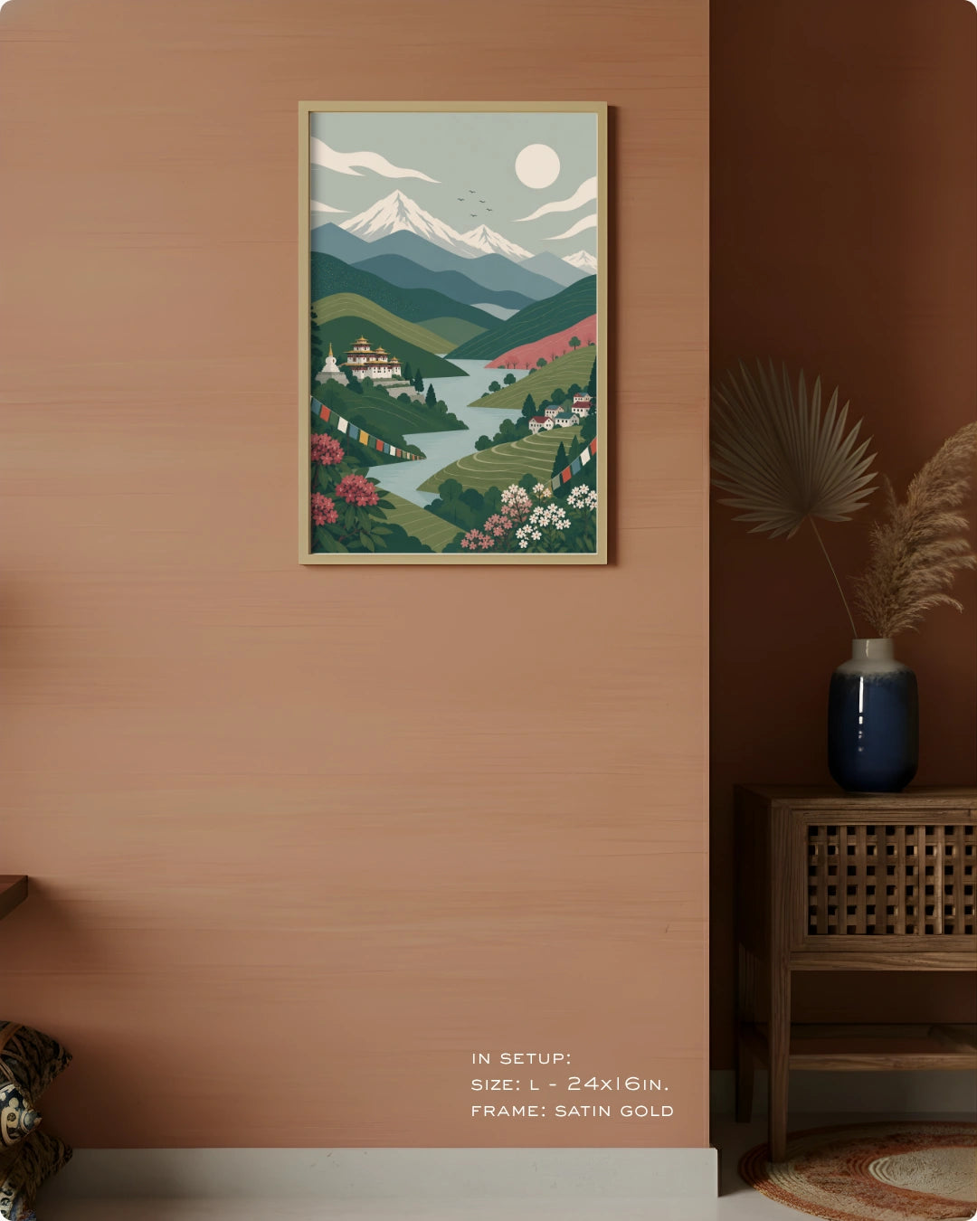 A framed art print depicting a stylized mountain landscape with a river, hills, and a moon, displayed on a peach-colored wall in a styled interior setting with a wooden side table and vase.