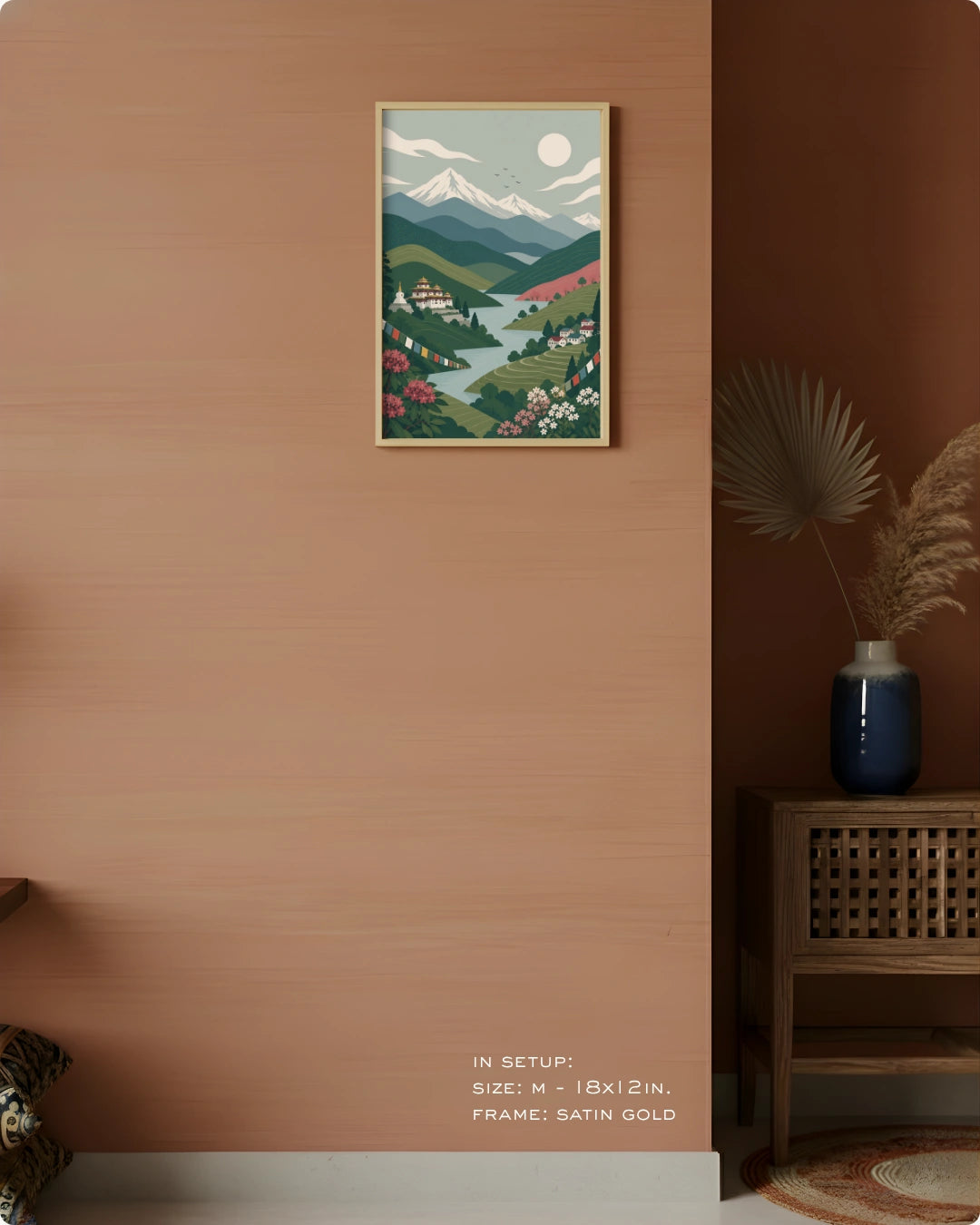 A framed art print depicting a stylized mountain and valley landscape hangs on a warm terracotta wall in a styled interior. The print shows green hills, pink flowers, a blue river, and white mountains under a pale sky.
