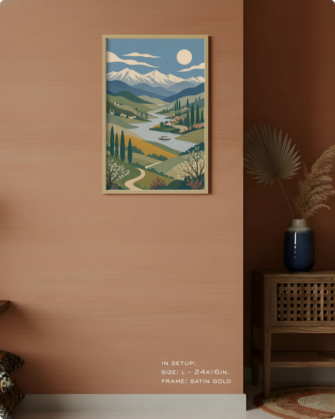 A framed landscape art print hangs on a warm peach-colored wall in a styled interior setting. The print depicts a stylized scenic valley with mountains, trees, a winding river, and a full moon.