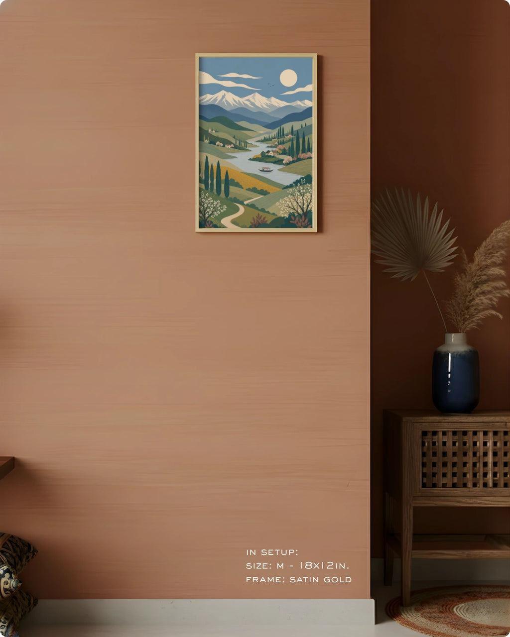 A framed landscape art print hangs on a warm terracotta wall in a styled interior. The print depicts a scenic valley with mountains, trees, a winding river, and a sun.