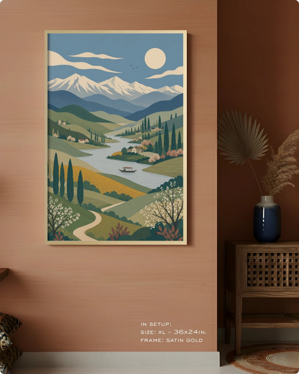 A framed landscape art print hangs on a warm terracotta wall in a styled interior. The artwork depicts a stylized valley scene with mountains, a winding river, trees, and a full moon.