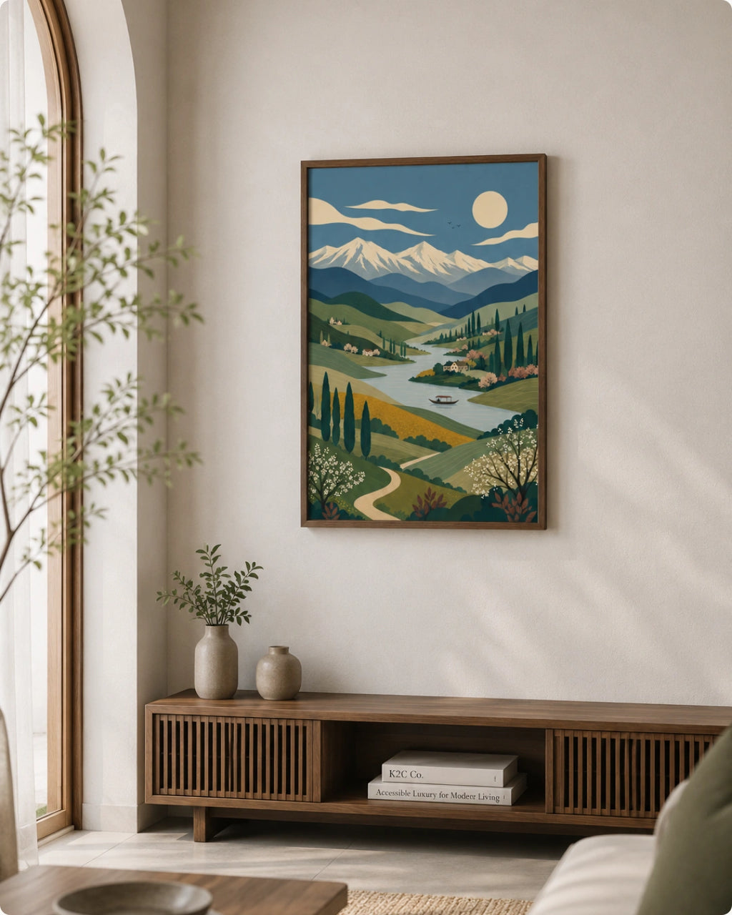 A framed landscape art print hangs on a light beige wall above a wooden console table in a modern living room setting. The print features a stylized illustration of a mountain valley with a winding river, trees, and a full moon.