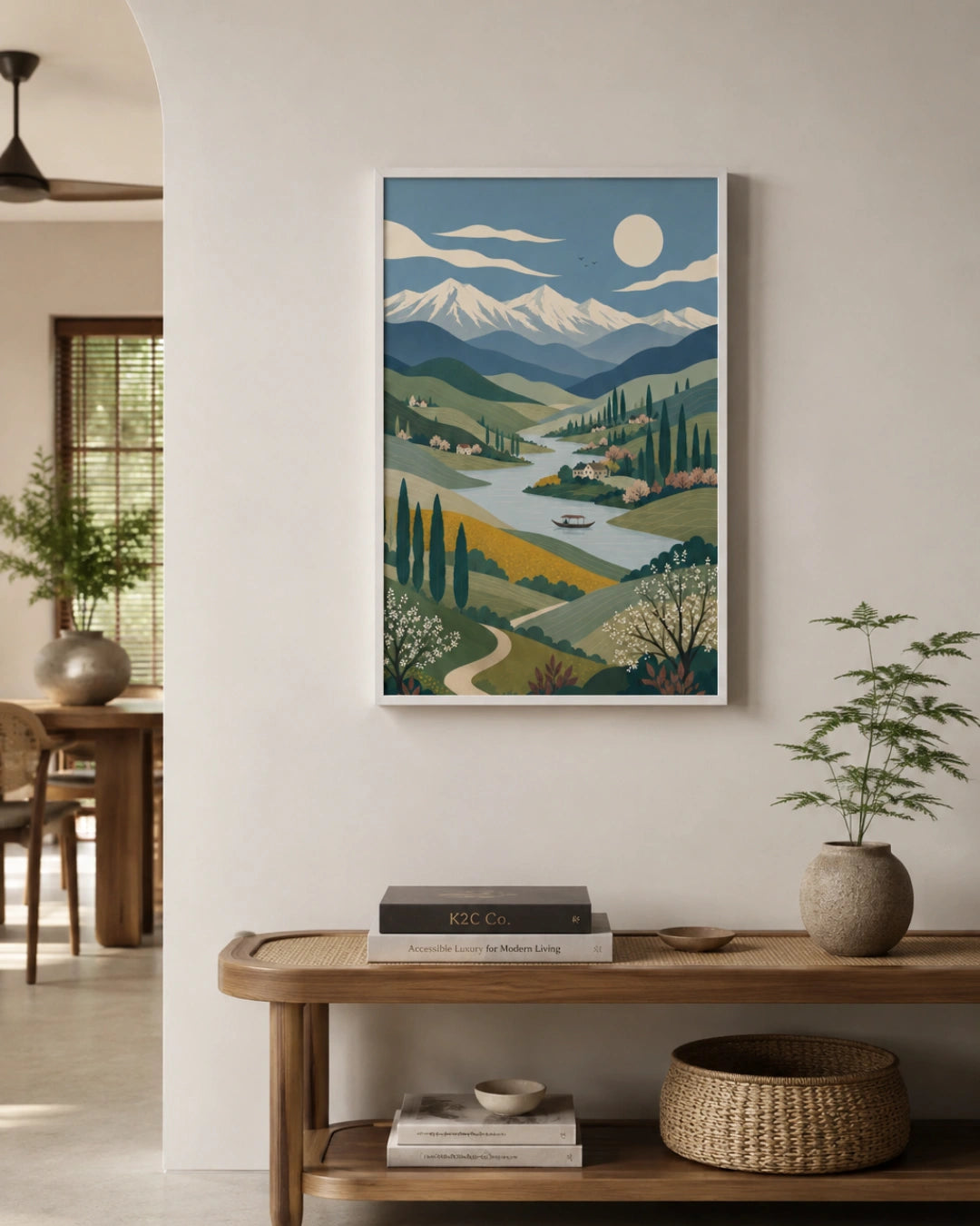 A framed art print featuring a stylized landscape with mountains, a river valley, and trees, displayed on a white wall above a wooden console table in a modern, neutral-toned interior.