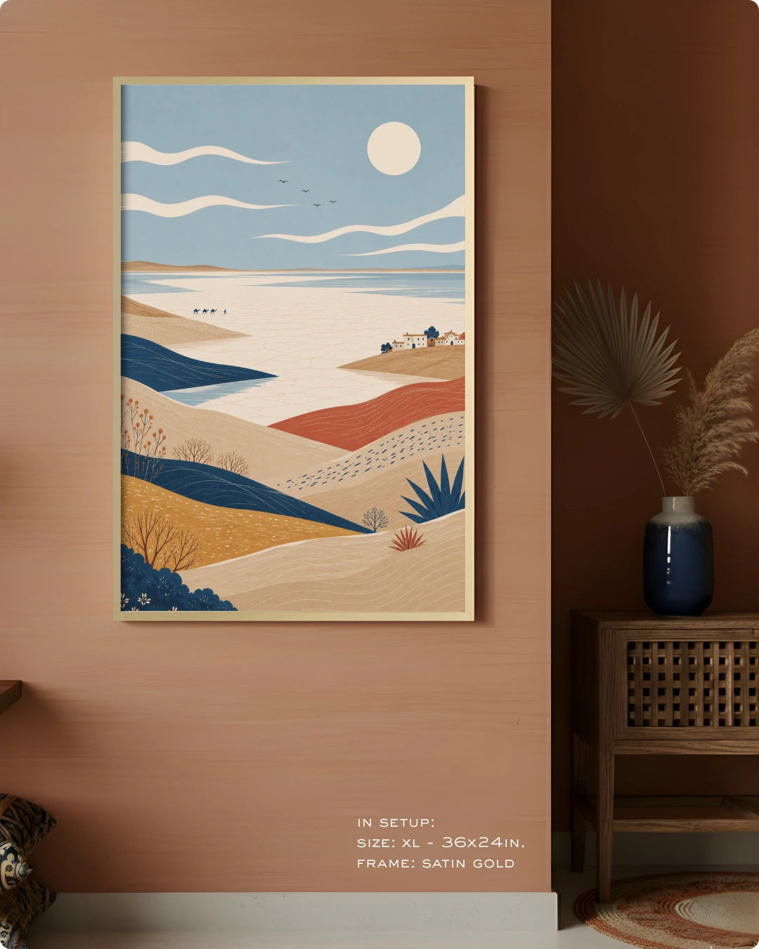A framed landscape art print hangs on a warm terracotta wall in a styled interior. The artwork depicts layered beige and blue hills, a pale blue sky with a white sun, and small coastal structures.