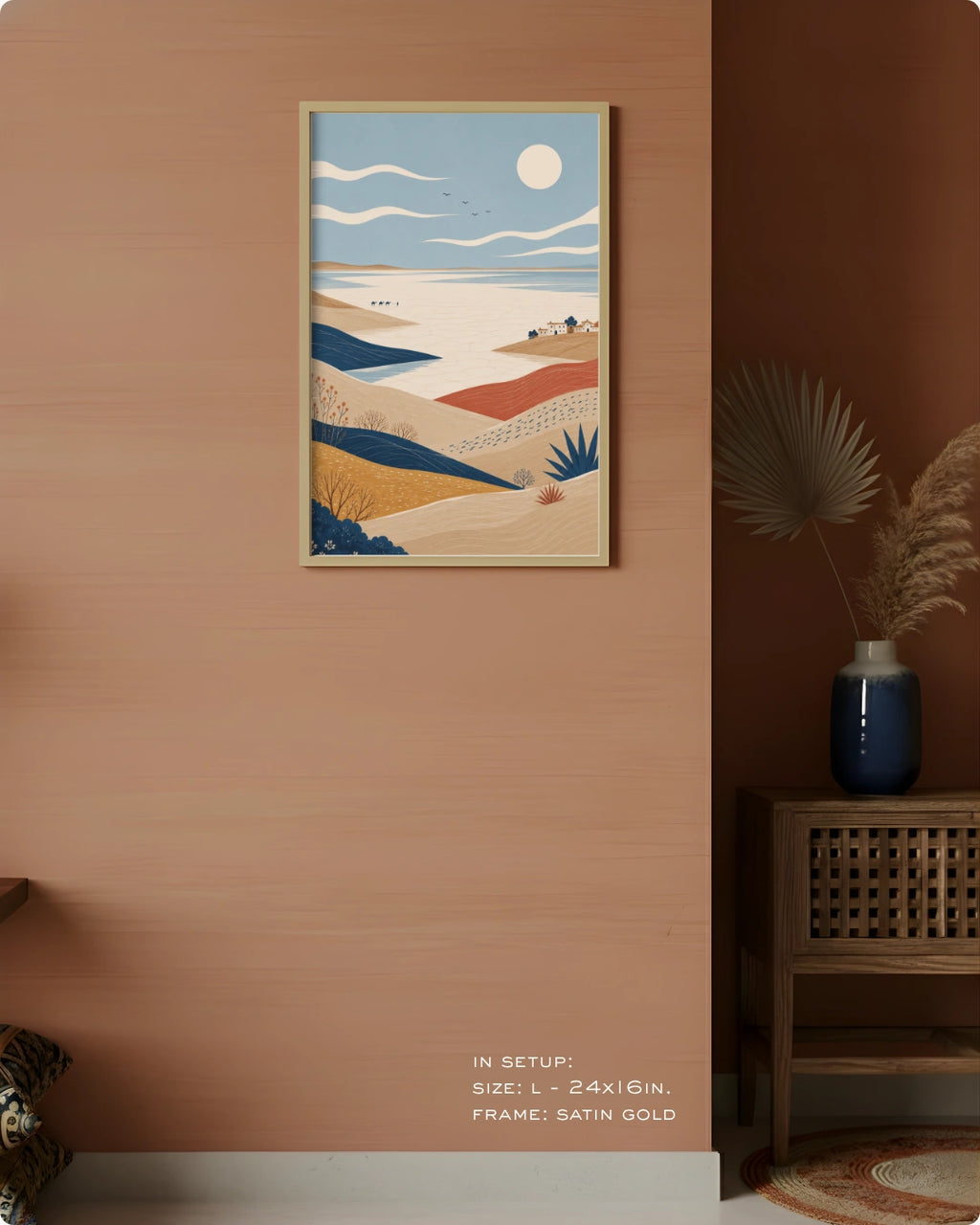 A framed vertical art print depicting a stylized desert landscape with dunes, a blue sea, and a sun, displayed on a warm terracotta wall in a styled interior setting.