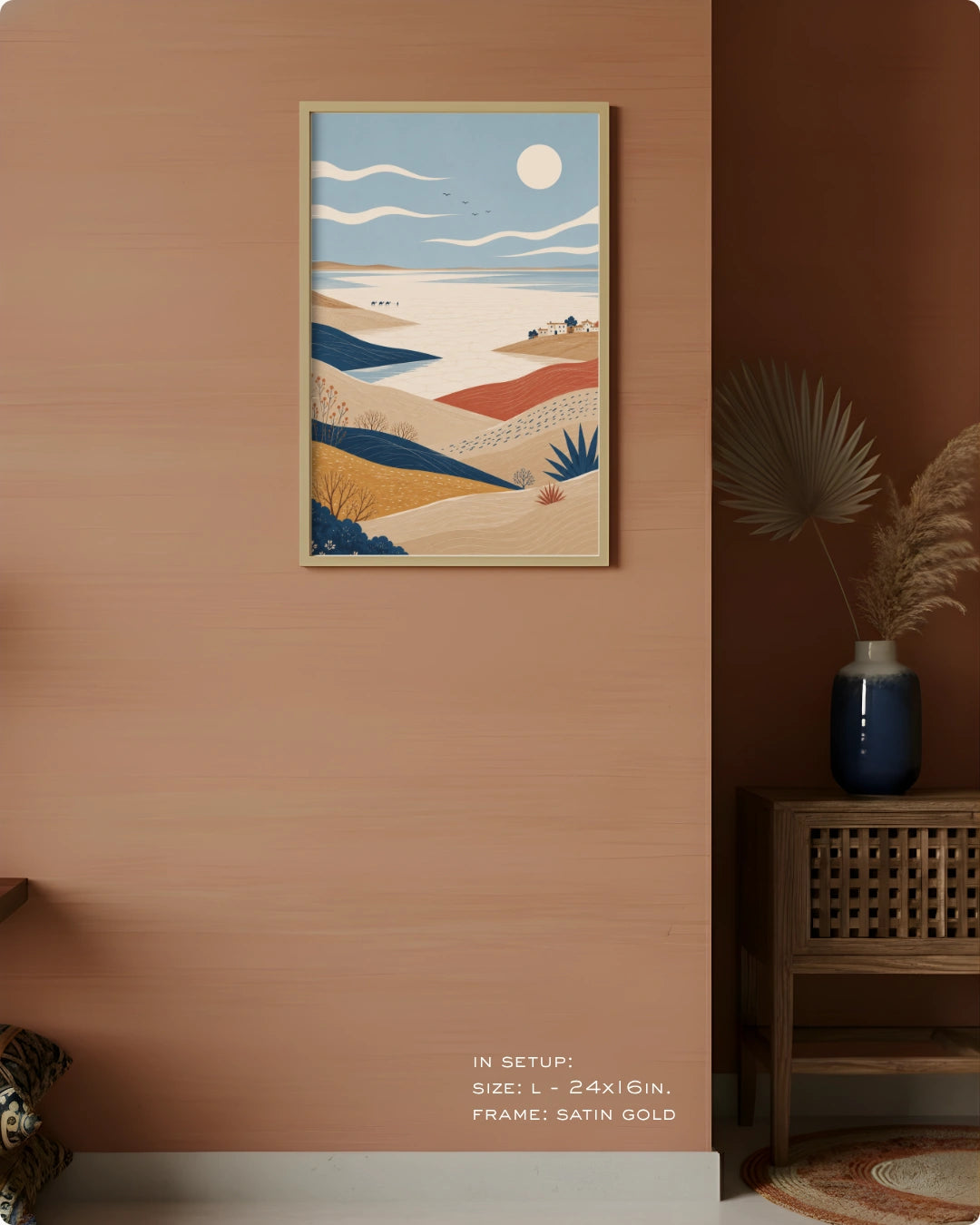 A framed vertical art print depicting a stylized desert landscape with dunes, a blue sea, and a sun, displayed on a warm terracotta wall in a styled interior setting.