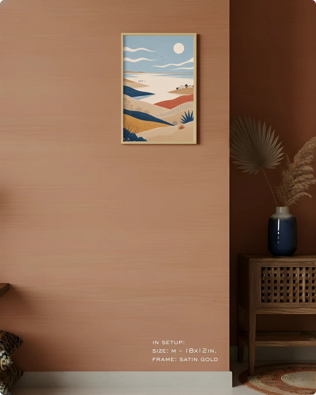 A framed art print featuring a stylized desert landscape with blue, beige, and orange tones, displayed on a warm terracotta wall in a styled interior setting.
