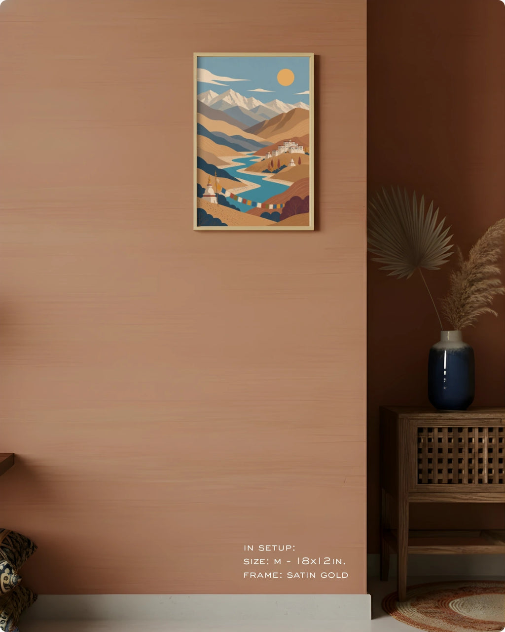A framed art print featuring a stylized landscape with mountains, a river, and a sun, displayed on a warm terracotta wall in a styled interior setting with a wooden cabinet and vase.