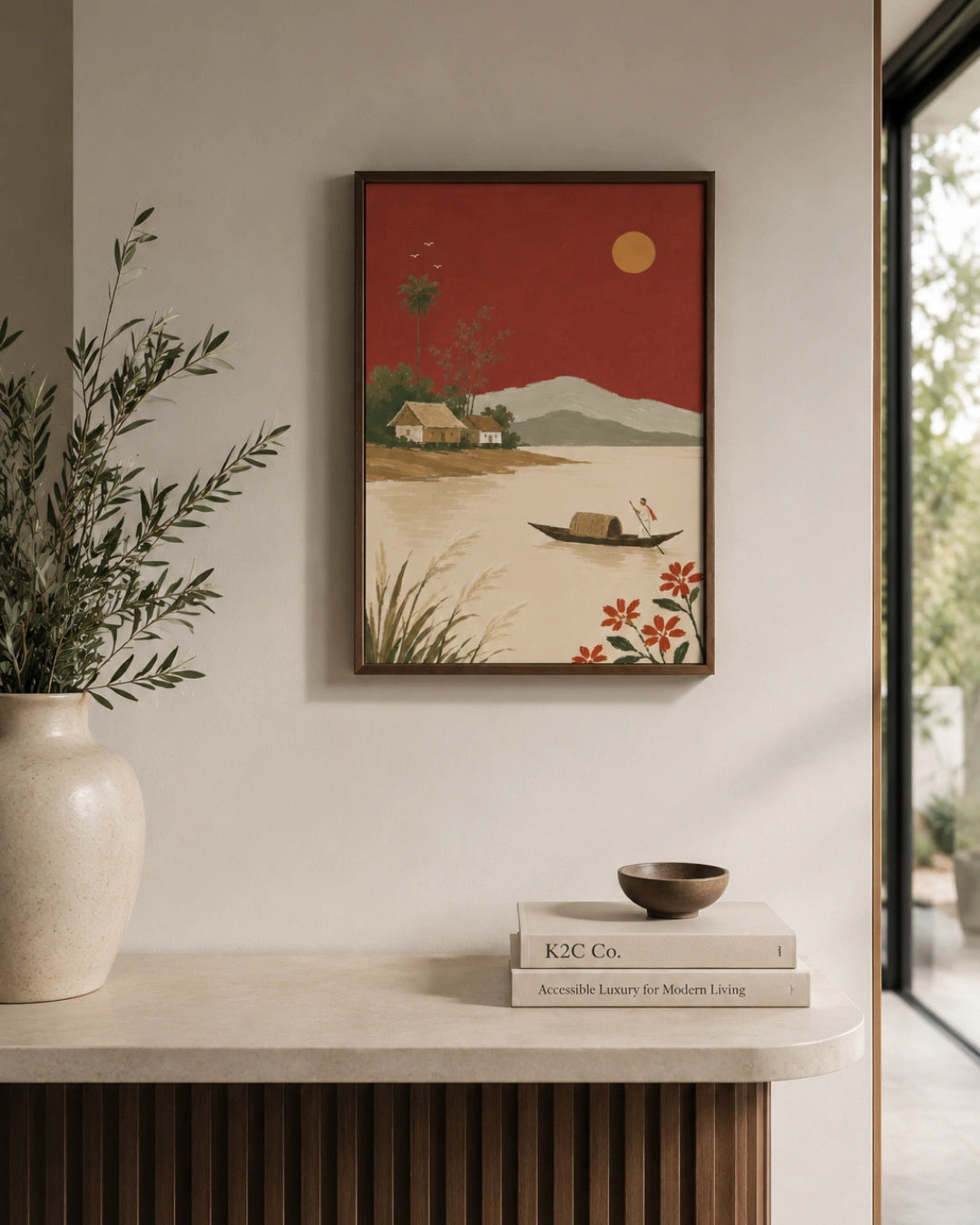 A framed art print depicting a serene landscape with a red sky, mountains, a boat, and flowers, styled on a beige wall above a console table with a vase and books.