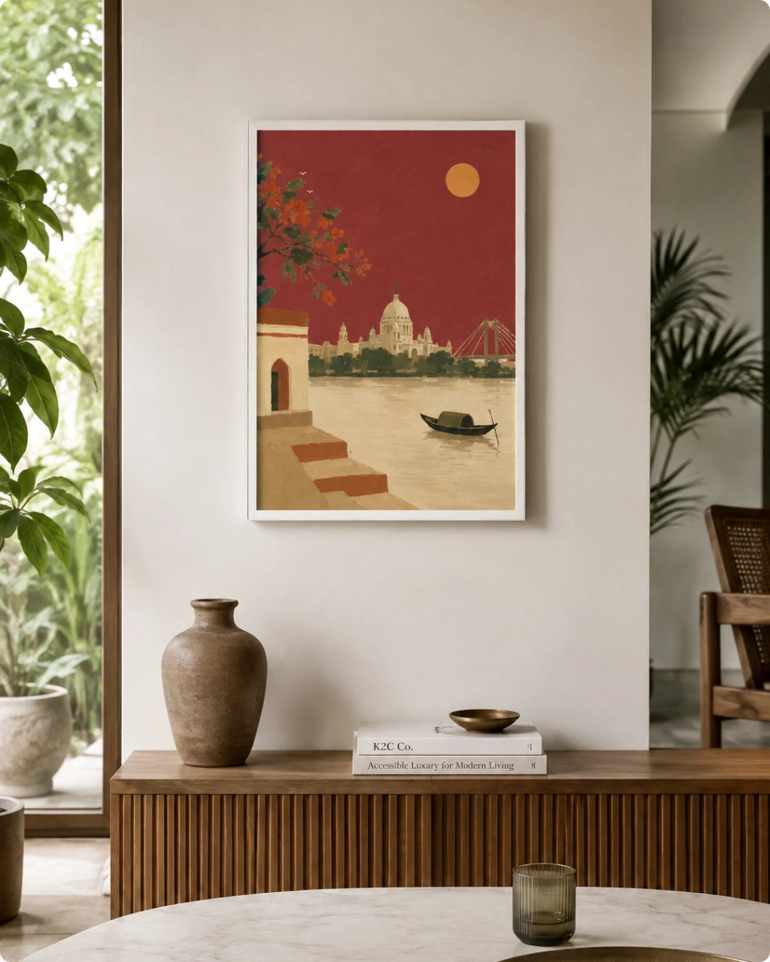 A framed art print featuring a stylized illustration of a river city at sunset, displayed on a beige wall in a modern, well-lit interior setting with a wooden console table, vase, and books.