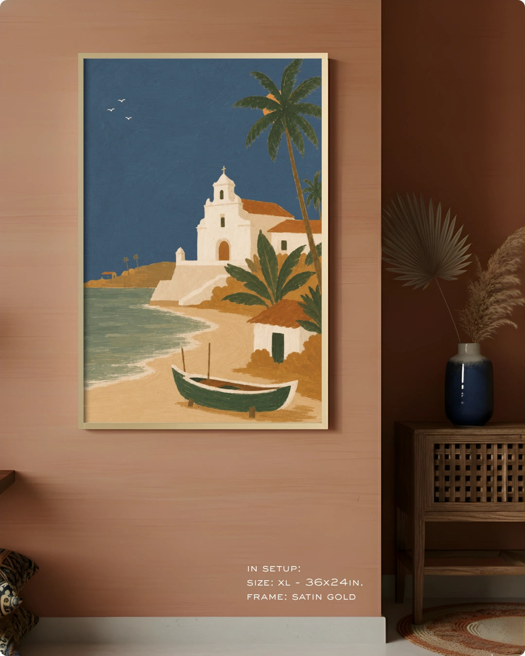 A framed coastal art print hangs on a warm terracotta wall in a styled interior. The artwork shows a white church on a sandy beach with palm trees, blue water, and a green boat.