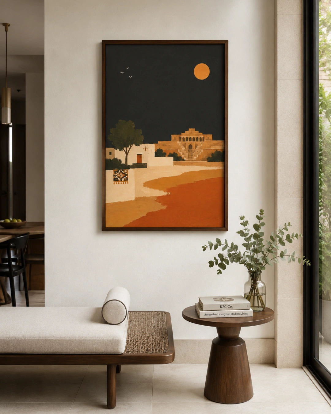 A framed art print depicting a stylized desert landscape with a golden sun, beige buildings, and a winding orange path under a dark night sky, displayed on a light beige wall in a modern, minimalist living room setting.