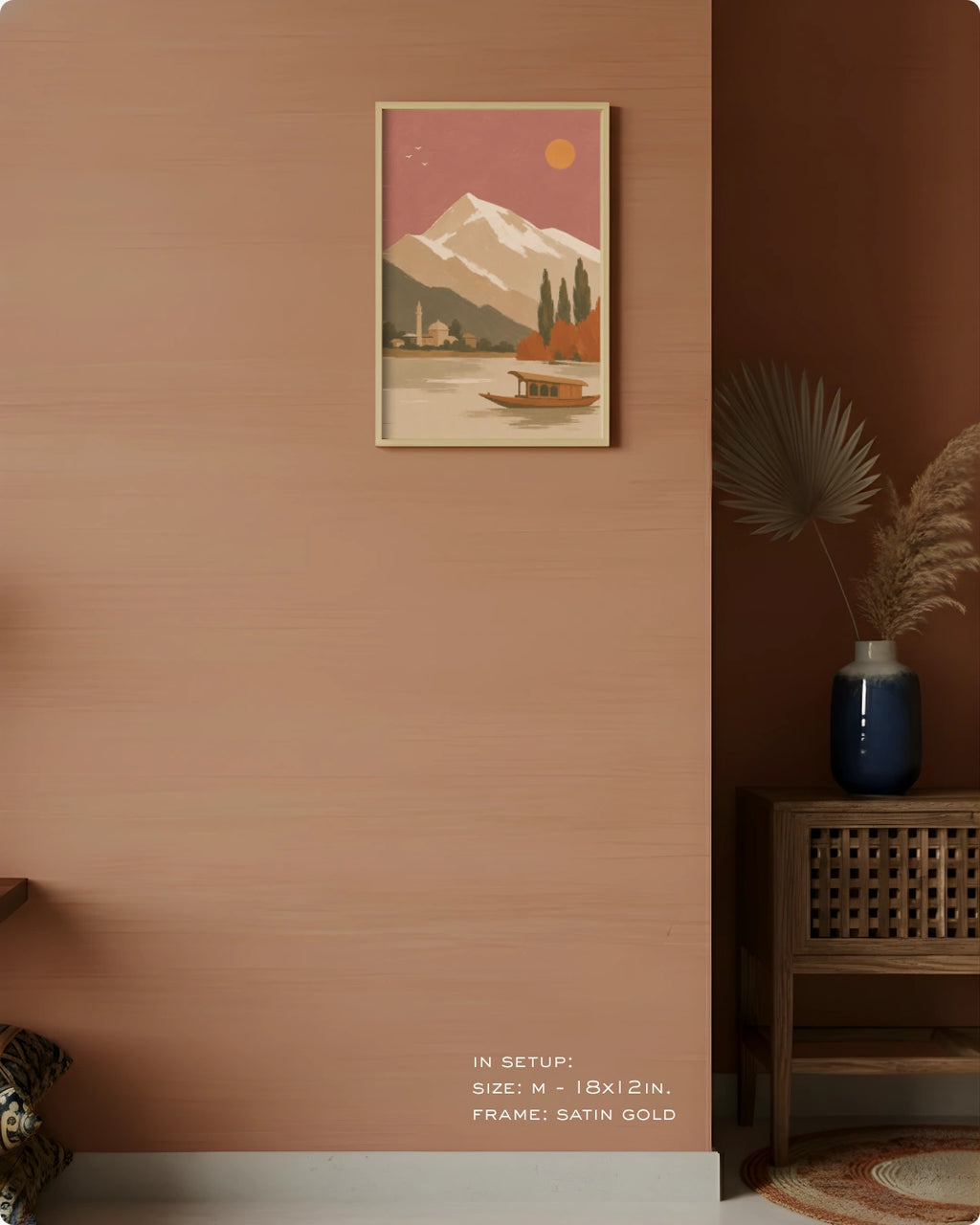 A framed art print depicting a mountain lake scene with a boat, hung on a terracotta-colored wall in a styled interior setting. The print features a pink sky, a snow-capped mountain, and a small boat on water, framed in satin gold.