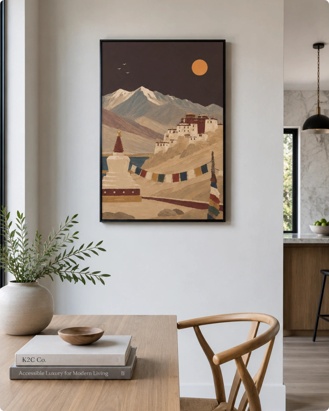 A framed art print depicting a stylized mountain landscape with a white stupa and traditional buildings under a yellow sun, displayed on a white wall in a modern dining room setting.