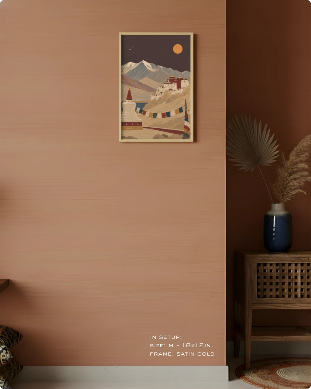A framed art print depicting a mountain monastery scene in warm earth tones, displayed on a terracotta-colored wall in a styled interior setting with a wooden cabinet, blue vase, and dried palm leaves.