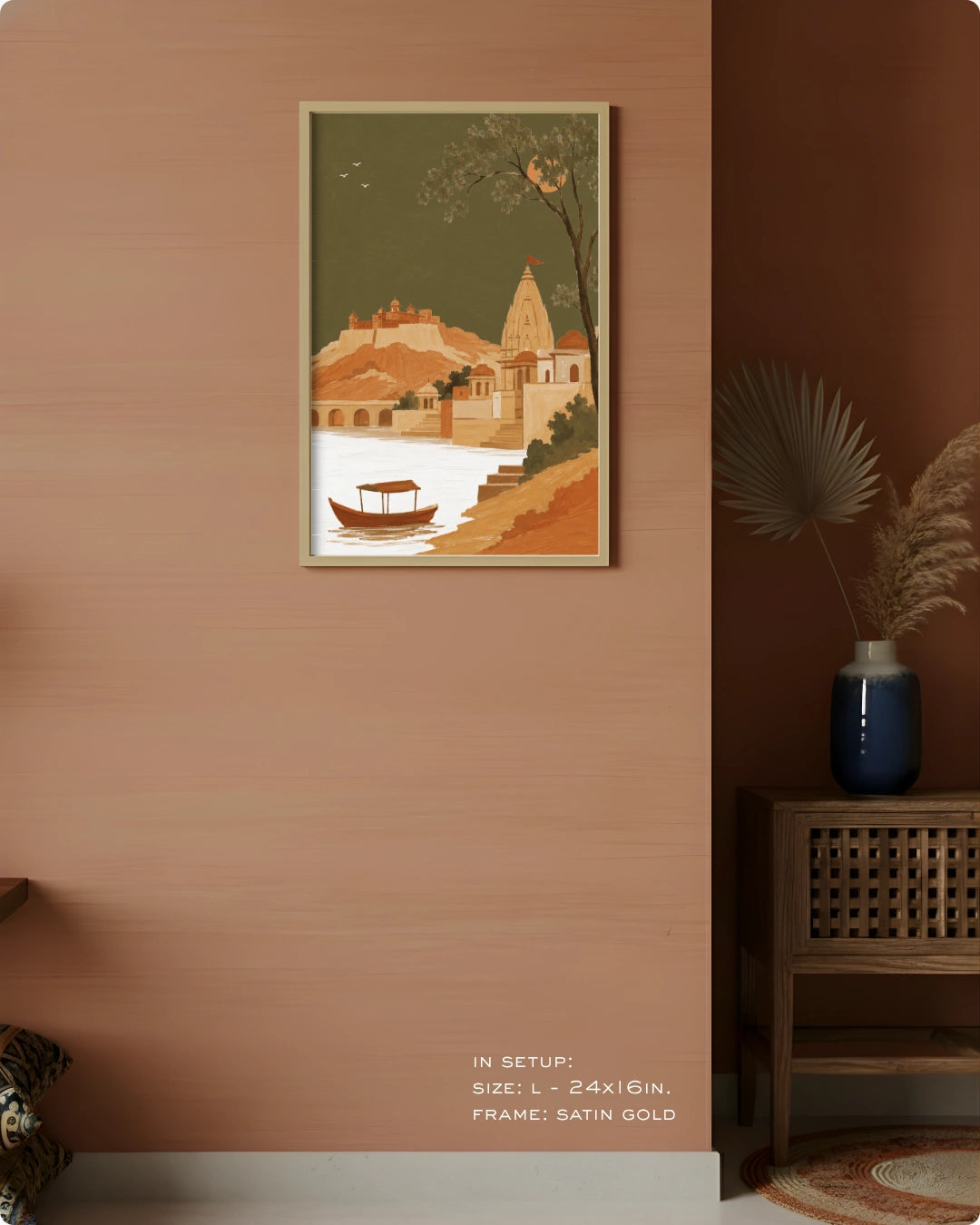 A framed art print depicting a scenic Indian coastal town with a boat on the water, displayed on a warm terracotta-colored wall in a styled room setting.