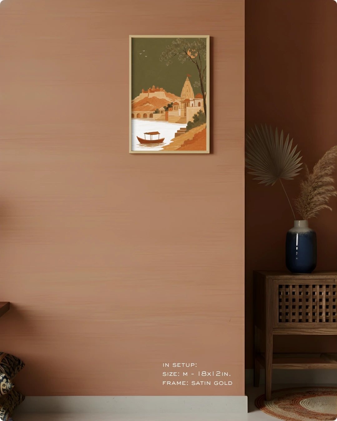 A framed vertical art print depicting a stylized village scene with a boat on a river, hung on a warm terracotta wall in a styled interior. The print has a muted green sky, orange-brown buildings, and a white river area, in a satin gold frame.