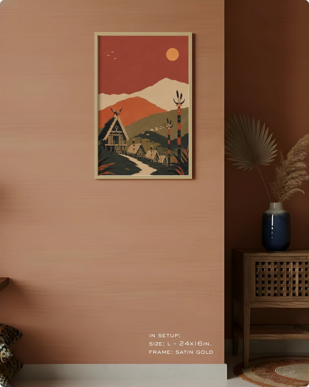 A framed art print featuring a retro-style mountain village scene in warm terracotta, cream, and forest green tones, displayed on a peach-colored wall in a styled interior setting with a wooden cabinet and dried palm in a blue vase.