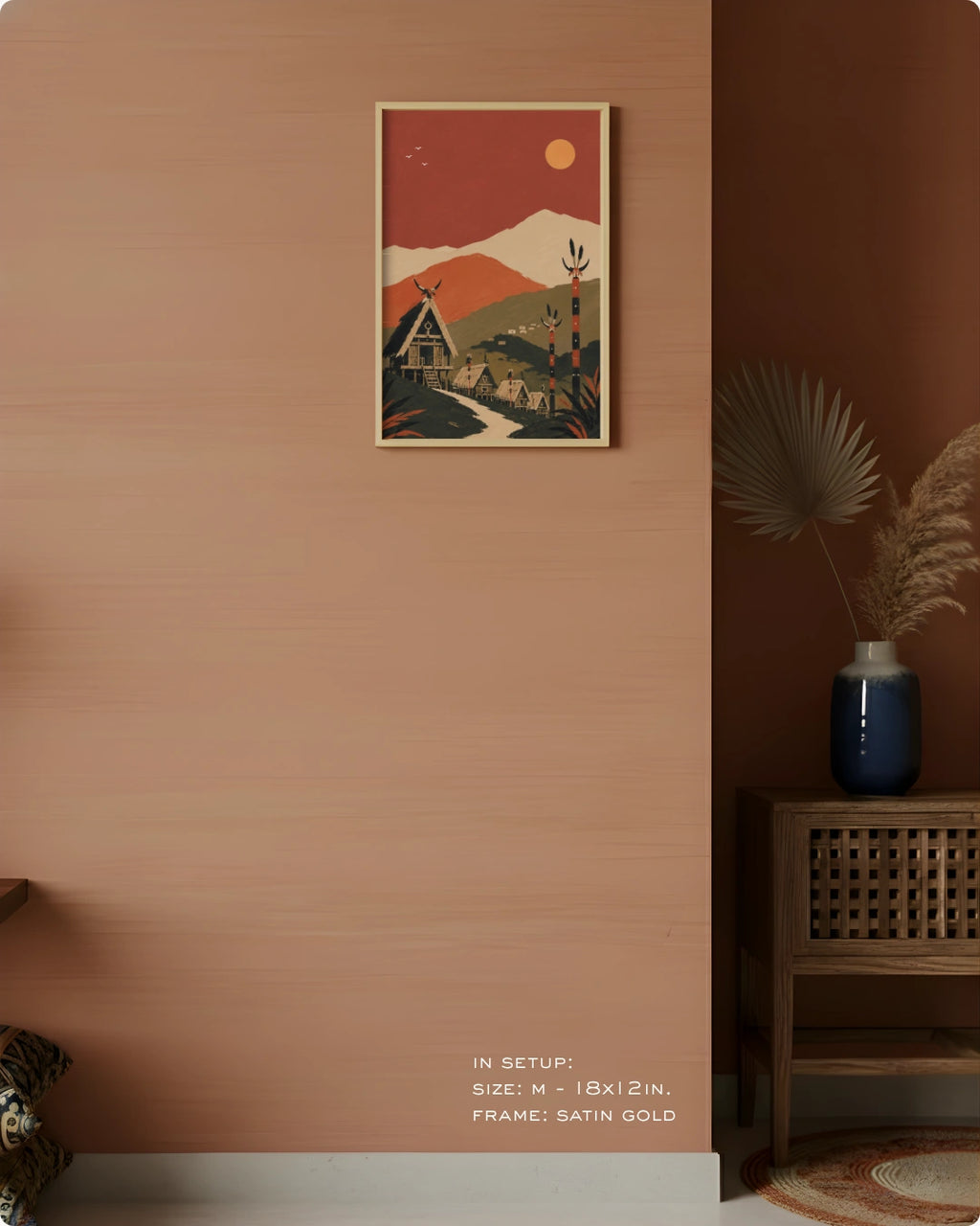 A framed art print featuring a retro-style mountain landscape with a house and deer, hung on a terracotta-colored wall in a styled interior setting with a wooden cabinet and dried pampas grass.