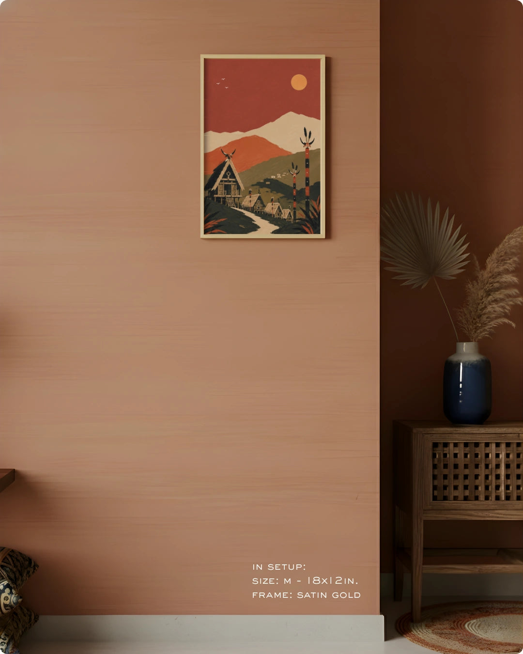 A framed art print featuring a retro-style mountain landscape with a house and deer, hung on a terracotta-colored wall in a styled interior setting with a wooden cabinet and dried pampas grass.