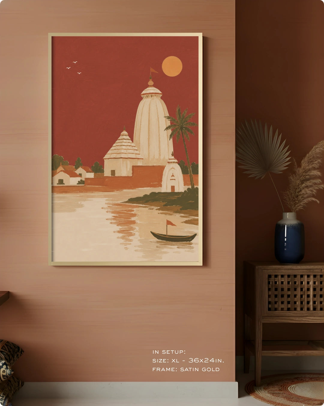 A framed art print depicting a stylized temple scene with a red sky, cream buildings, palm trees, and water, displayed on a warm brown wall in a styled interior setting.