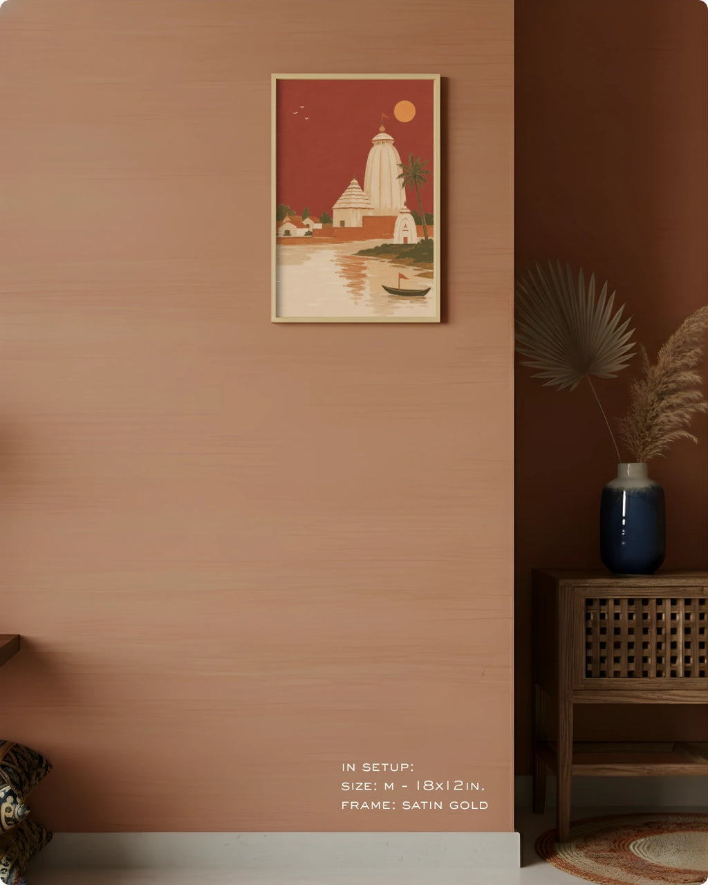 A framed art print depicting a temple and water scene in warm red, cream, and gold tones, hung on a terracotta-colored wall in a styled interior setting with a wooden cabinet, blue vase, and dried palm leaves.