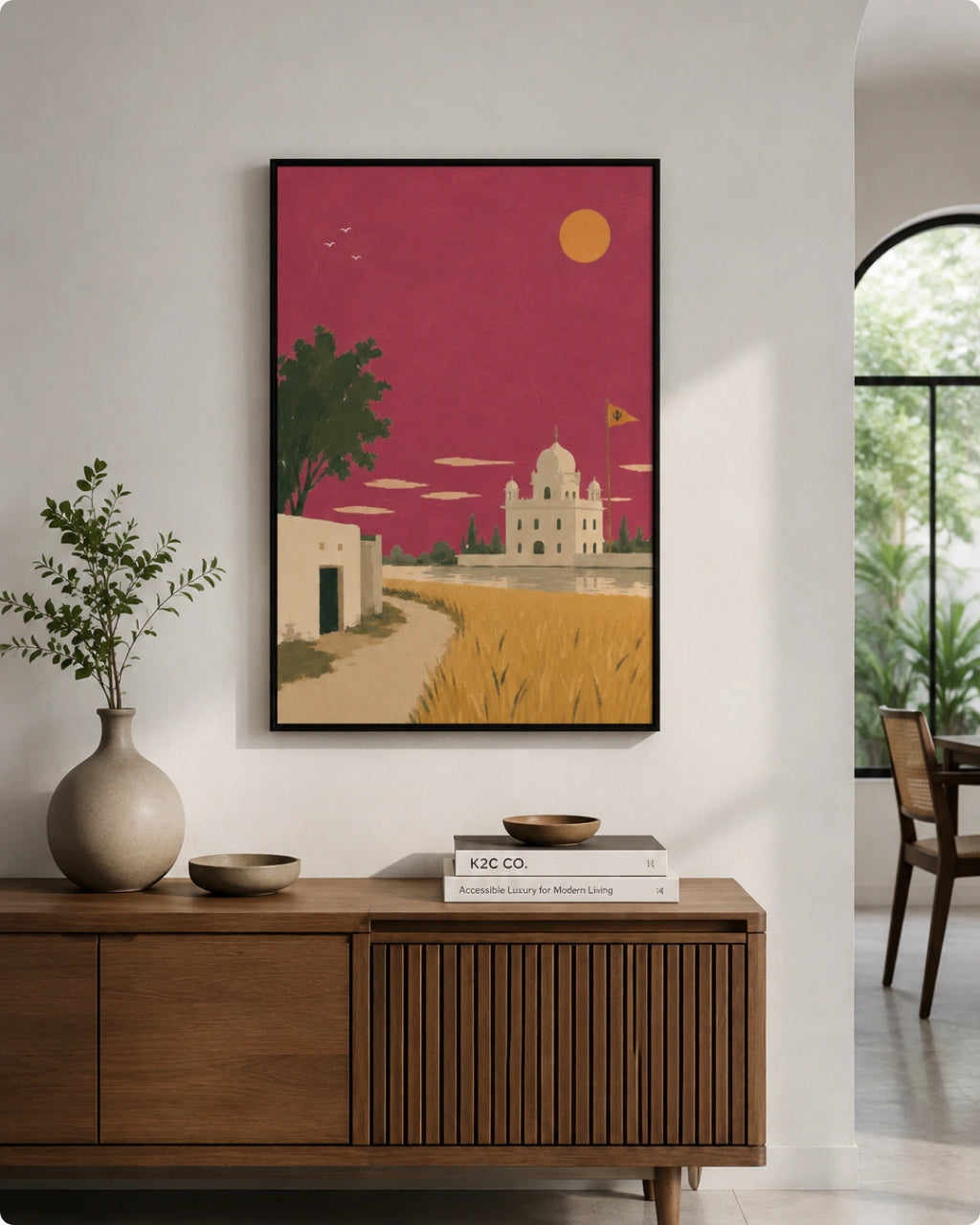 A framed art print featuring a stylized desert landscape with a white domed building, golden fields, and a magenta sky, displayed on a white wall above a wooden console table in a modern interior setting.