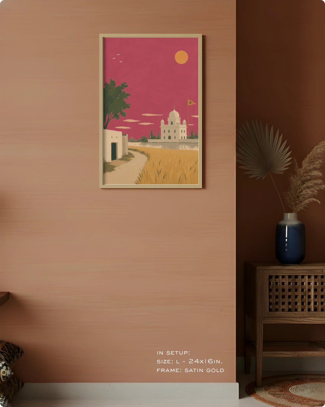 A framed art print featuring a stylized desert landscape with a white domed building, golden fields, and a pink sky, displayed on a peach-colored wall in a styled interior setting.