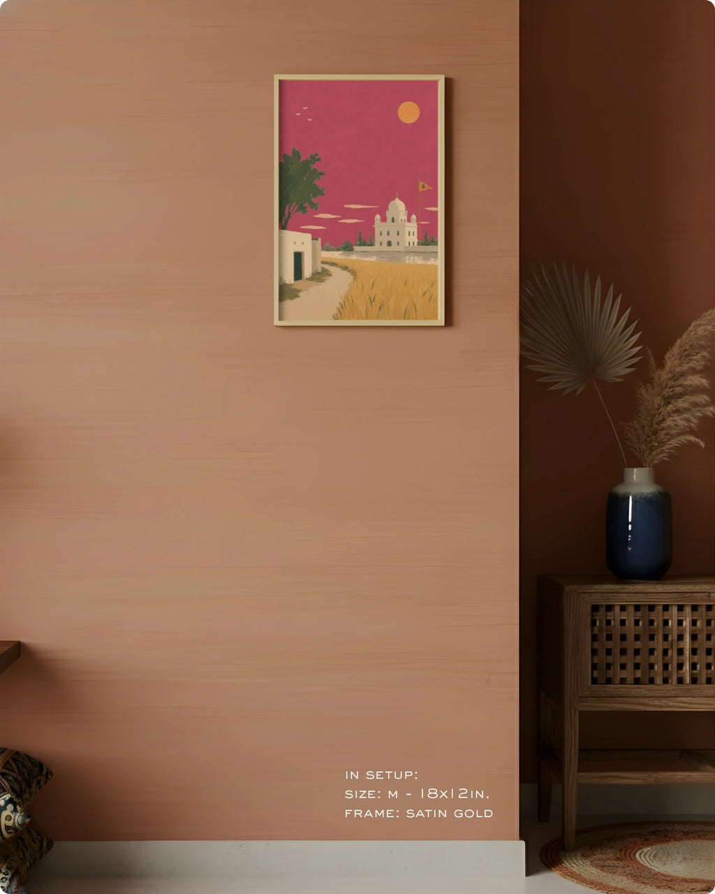 A framed art print depicting a stylized desert landscape with a white domed building under a pink sky, hung on a warm terracotta wall in a styled interior setting with a wooden side table, blue vase, and dried palm leaves.