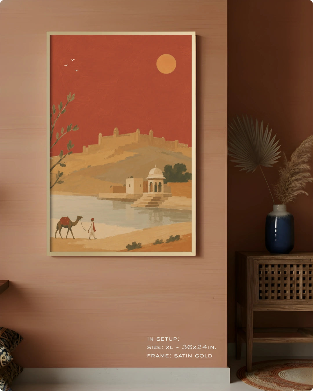 A framed art print depicting a desert landscape with a camel, a person, a domed building by a river, and a golden sun on a terracotta background, displayed on a warm terracotta wall in a styled interior setting.